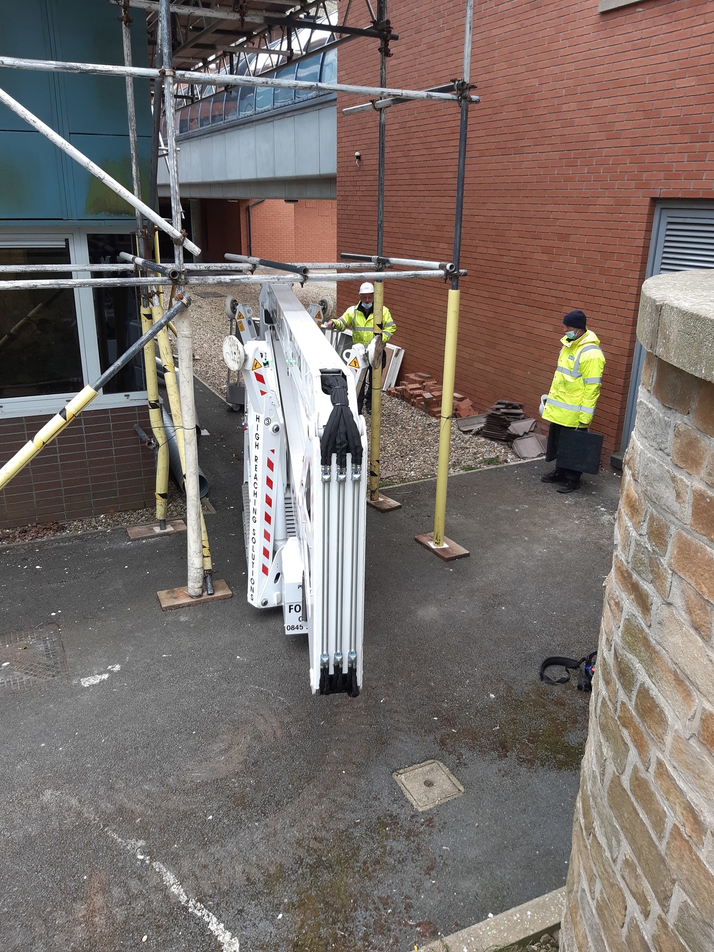 Lottie, High Reaching Solutions 20m Lithium hybrid tracked spider lift cherrypicker, on narrow tracked setting, moving through Scaffolding to exit the worksite in Scarborough, North Yorkshire.