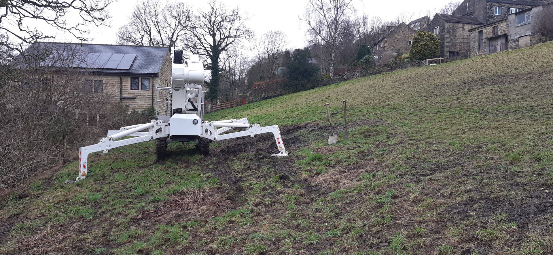Selina, High Reaching Solutions 22m lithium hybrid tracked spider access platform, set-up on steep grass bank with two legs high up the slope and two lower down the slope. The bank had to be dug-out for the feet to be level to avoid Selina slipping down the bank.